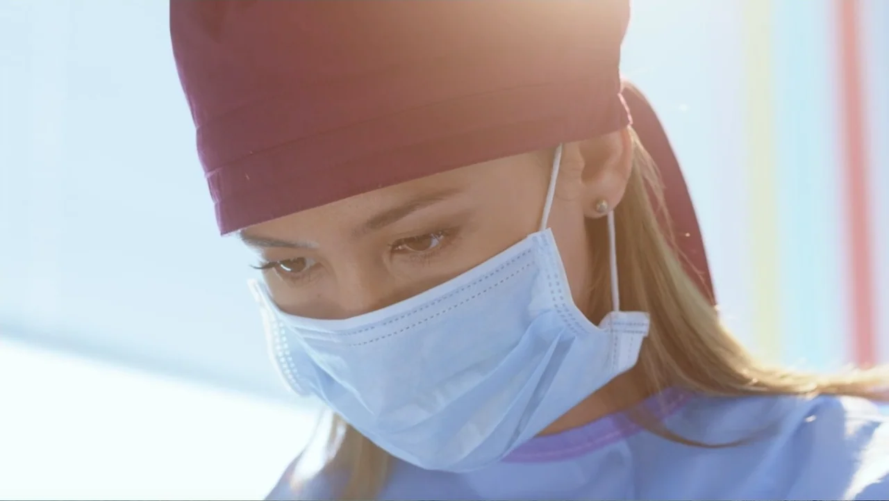Christmas with a Prince (2018) movie still: closeup — A clinical medical environment with bright overhead lighting. A female surgeon looks down…