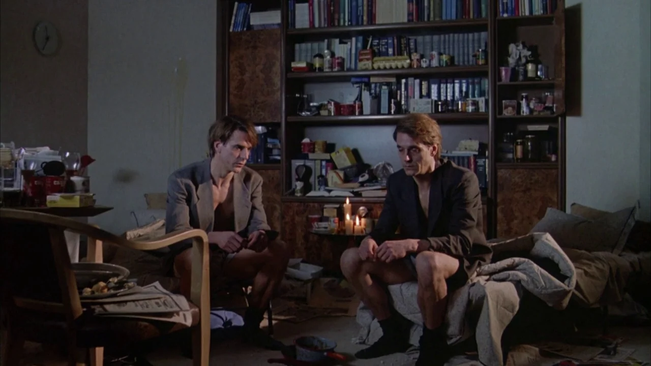 Dead Ringers (1988) movie still: medium — A cluttered apartment room contains a large filled bookshelf and two men sitting nearby. The men are…