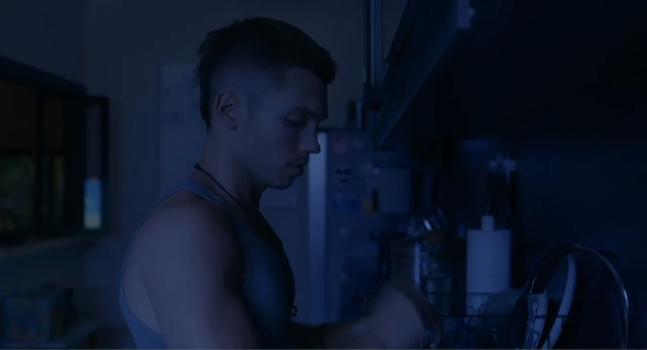 Straight (2024) movie still: medium — A dimly lit home kitchen at night with blue tones and muted shadows. A young man in a grey tank top stands…