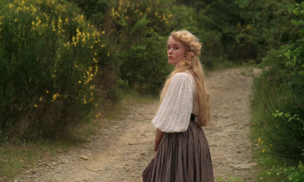 A Room With A View (1985) movie still: medium — A rural dirt path winds through lush greenery and yellow flowering bushes. A young woman in a…