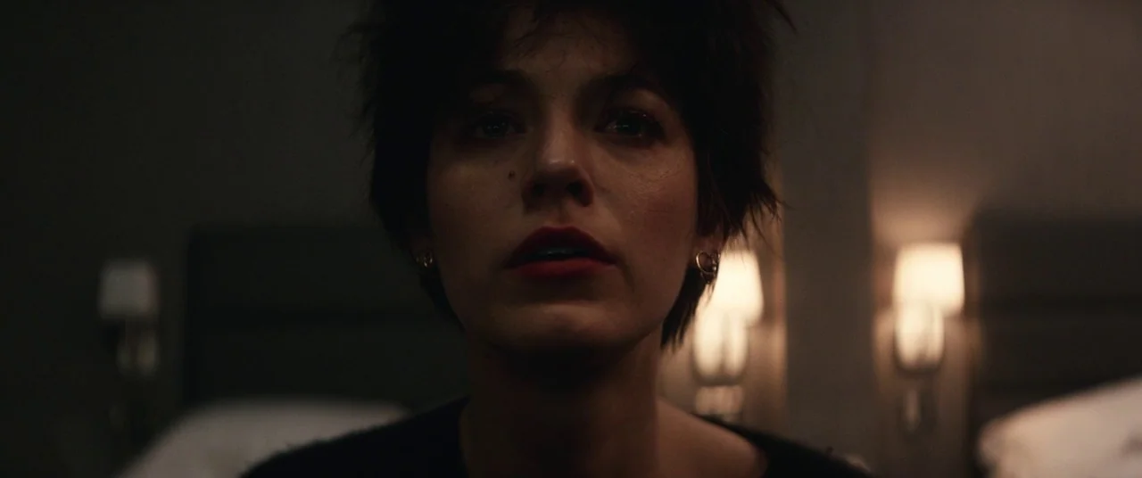 The Rhythm Section (2020) movie still: closeup — A dimly lit room features two blurred bedside lamps glowing against a dark wall. A woman looks…