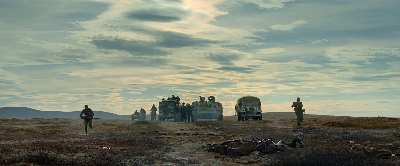 Sisu (2022) movie still: wide — A group of soldiers and military vehicles traverse a vast, barren tundra landscape. In the foreground, a dead horse…