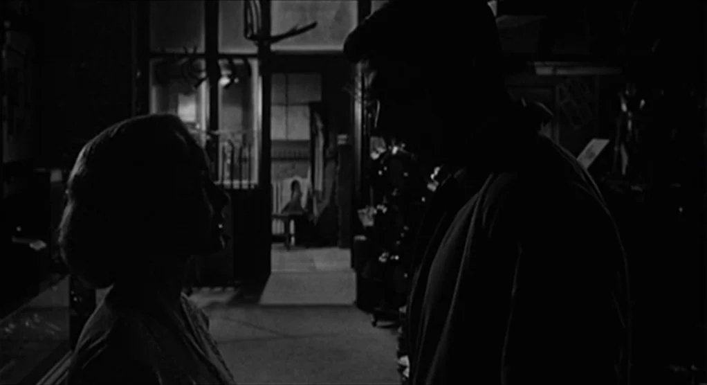 Psycho movie still: medium — The interior of a darkened storefront features silhouettes of two figures in conversation. Light streams through a…