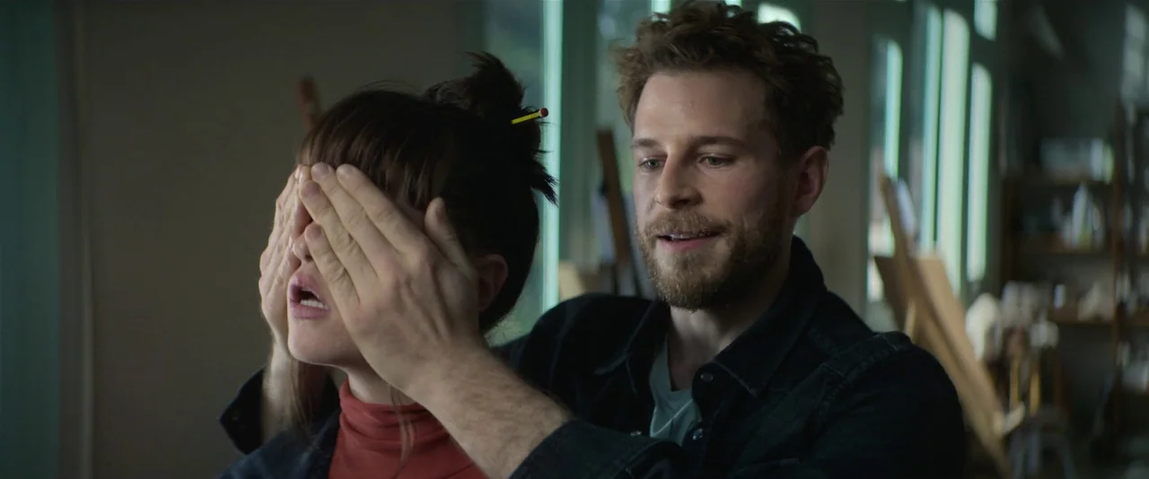 Crazy About Her (2021) movie still: medium — An art studio features wooden easels and large windows in the background. A man standing behind a woman…
