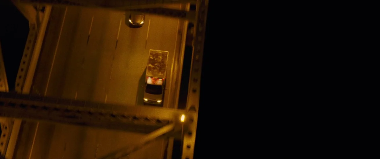 Synchronic (2019) movie still: establishing — A top-down view shows an elevated highway crossing a dark structure at night. A lone white delivery…