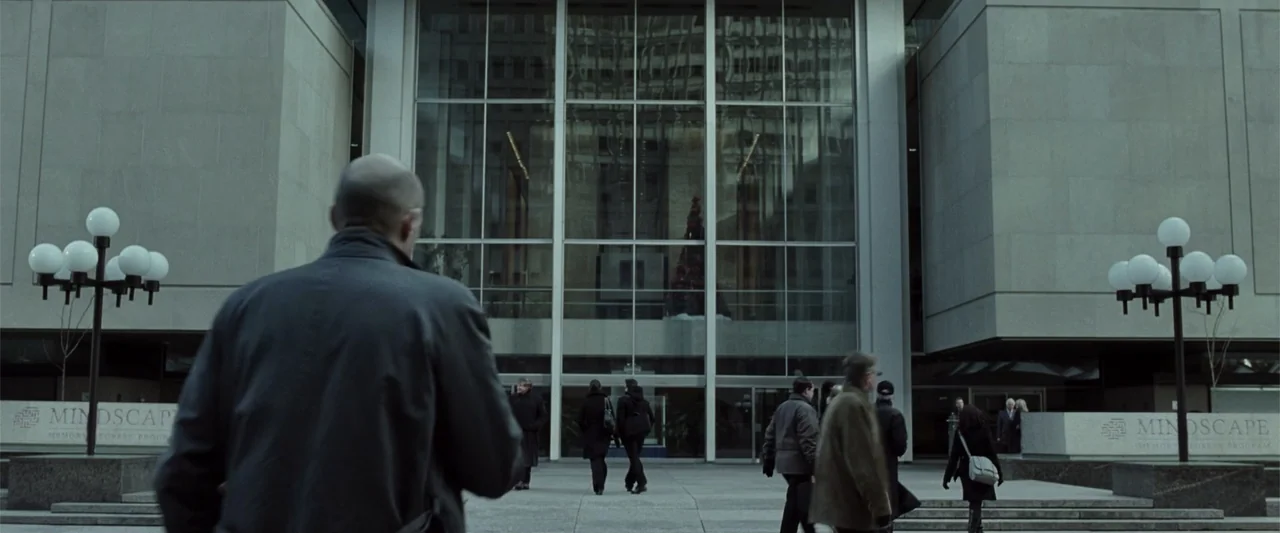Anna (2013) movie still: medium — A modern office plaza features a large glass building entrance with stone facades. A man in a dark coat walks…