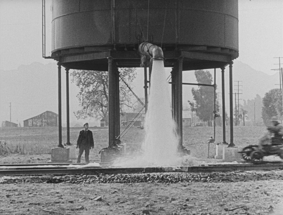 Sherlock Jr. (1924) movie still: wide — A rural railway area features a massive metal water tank on thick industrial stilts. A man stands on the…