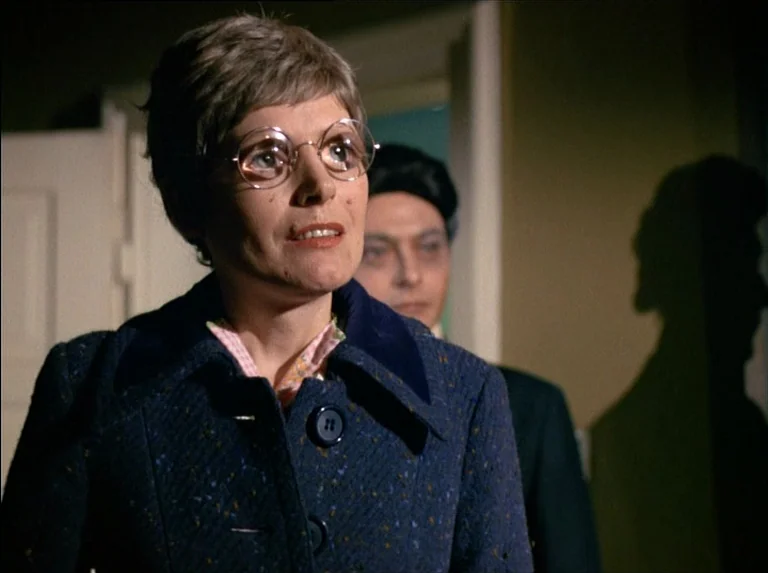Satan’s Brew (1976) movie still: medium — A dimly lit indoor space with a plain wall and a door frame in background. A woman in glasses and a dark…