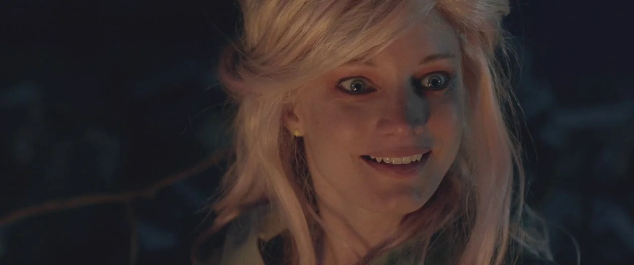 Turbo Kid (2015) movie still: closeup — A dark, out-of-focus forest setting at night serves as the backdrop. A young woman with blonde hair stares…
