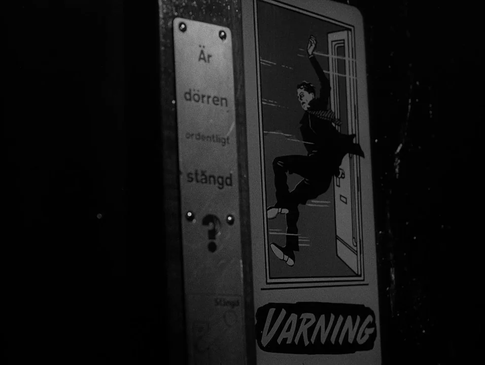 Dreams (1955) movie still: closeup — A metal warning sign is mounted to a dark wall inside a vehicle. The sign features a stylized illustration of a…