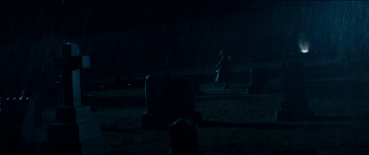 El Chicano (2018) movie still: wide — A dark, rain-swept graveyard features several stone monuments under a night sky. A lone figure in a long coat…