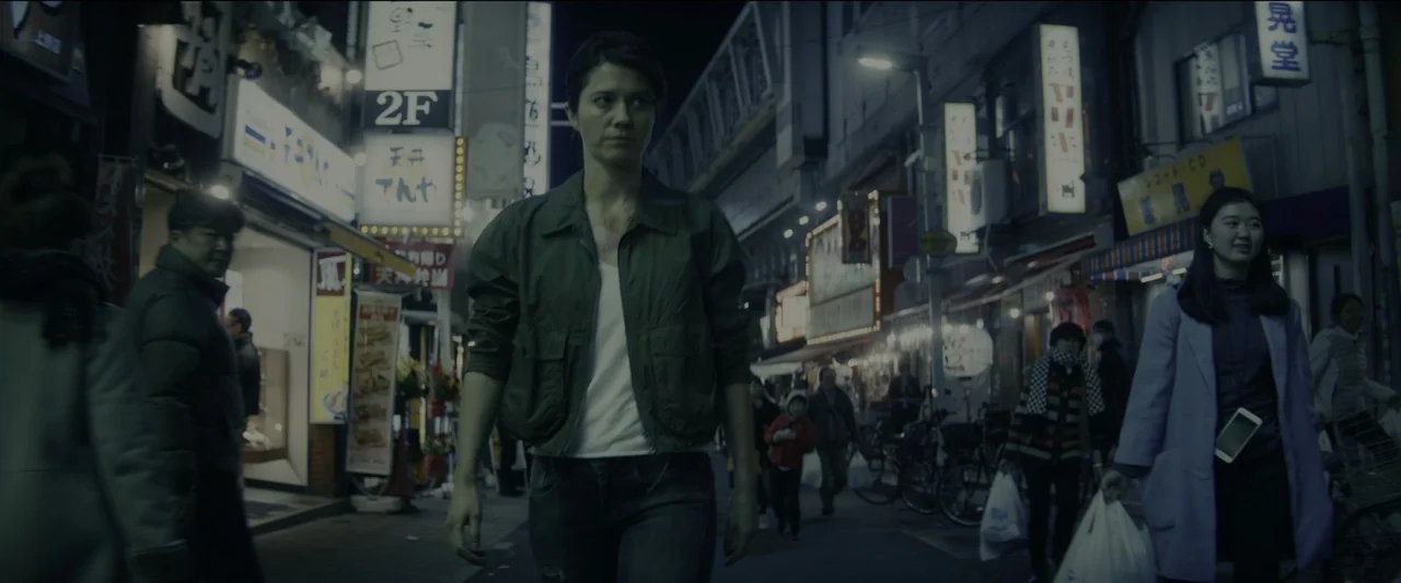 Kate (2021) movie still: medium — A crowded Japanese city street at night is lined with illuminated neon store signs. A woman in an olive jacket…