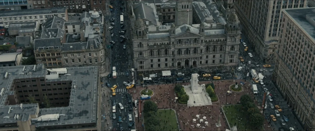 World War Z (2013) movie still: establishing — A dense urban plaza features an ornate stone monument surrounded by pedestrian walkways and…