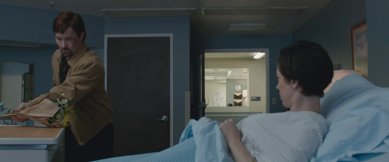 The Gift (2015) movie still: medium — A quiet hospital room with light blue walls and a closed door beside the entrance. A man in a tan jacket…