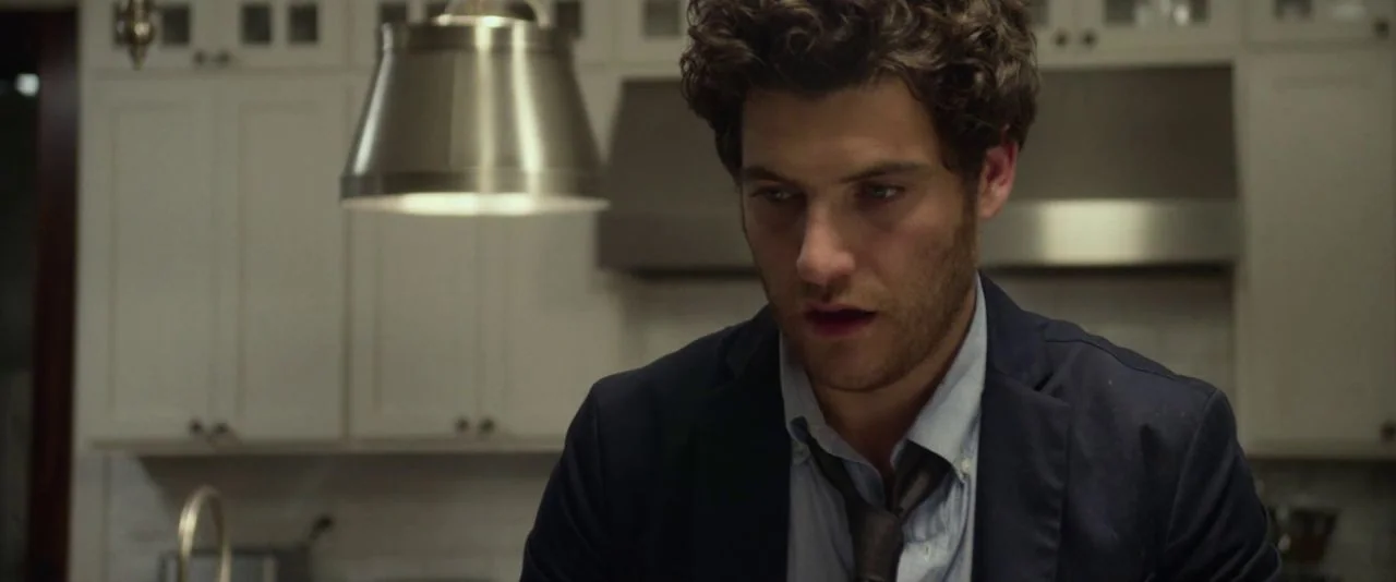Night Owls (2015) movie still: medium — A modern white cabinetry background with a brushed metal pendant light overhead. A man in a suit looks down…