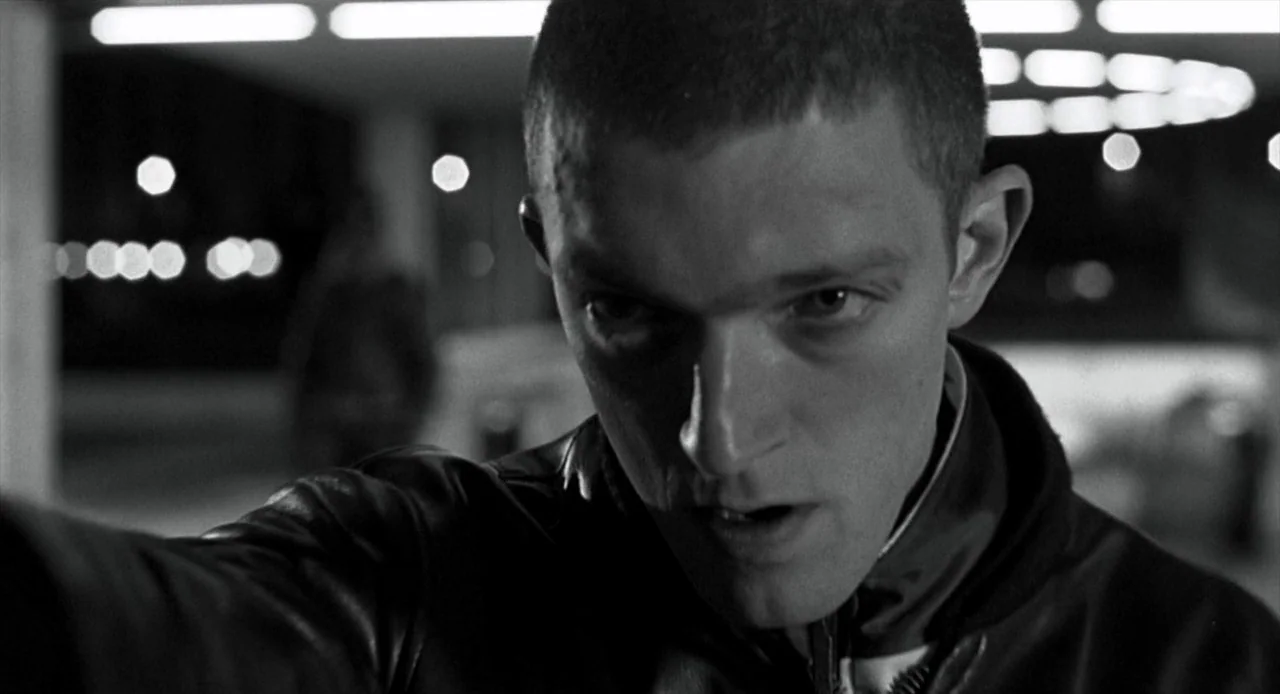La Haine (1995) movie still: closeup — A dimly lit concrete parking garage structure at night with blurred background lights. A young man with a…