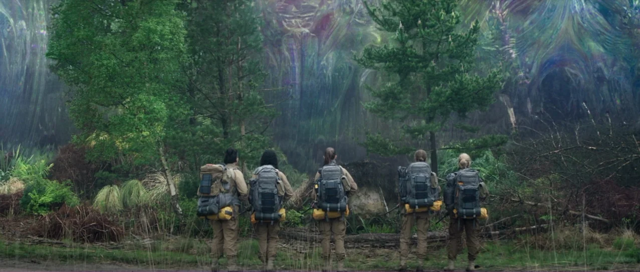 Annihilation (2018) movie still: wide — Five explorers stand in a line facing an iridescent, shimmering atmospheric anomaly in a forest. The anomaly…