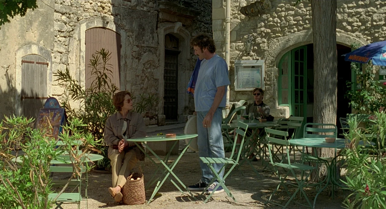 Swimming Pool (2003) movie still: medium — A stone-walled building with a green door frames a sunlit outdoor cafe terrace. A woman sits at a round…