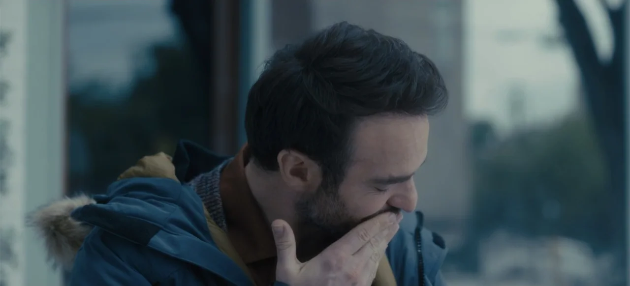 Merv (2025) movie still: closeup — A man stands outdoors near a window on a gray day. He covers his mouth with his hand while looking down.