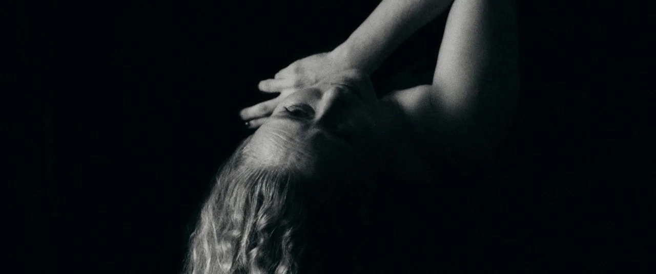 Saint Maud (2019) movie still: closeup — A dark, featureless space obscures all environmental details beyond the subject. A woman leans her head…