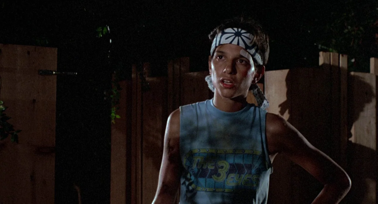 The Karate Kid (1984) movie still: medium — A dimly lit outdoor space at night features a plain wooden fence in background. A young male standing in…