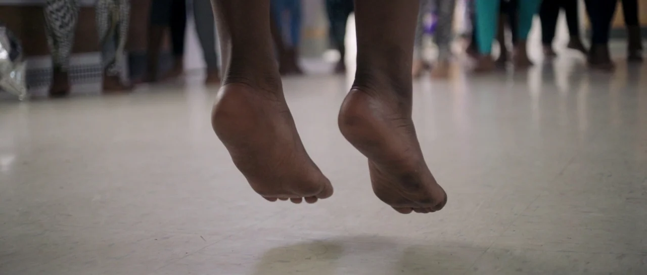 The Fits (2016) movie still: closeup — The frame focuses on a pair of dark-skinned feet suspended above a speckled linoleum floor. Several blurred…