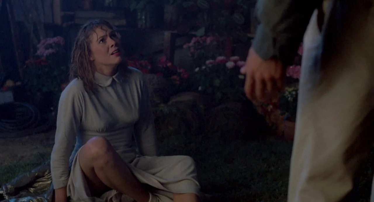 Peggy Sue Got Married (1986) movie still: medium — A dark outdoor space with grass and scattered flower pots under night lighting. A woman in a grey…