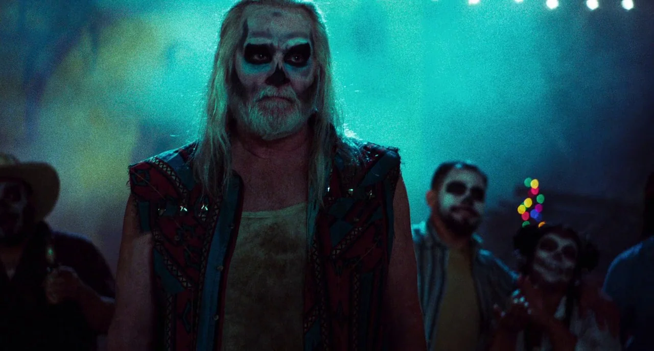 3 From Hell (2019) movie still: medium — A dimly lit room populated by people wearing skull makeup at a social gathering. An older man with long…