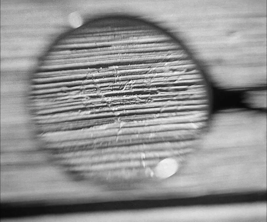 M (1931) movie still: closeup — A close-up of a circular textured surface embedded within a flat board. Fine parallel grooves run across the…