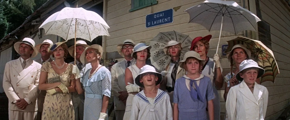 Papillon (1973) movie still: wide — A group of people in 1930s attire stand before a wooden wall. They hold umbrellas and look toward an object…