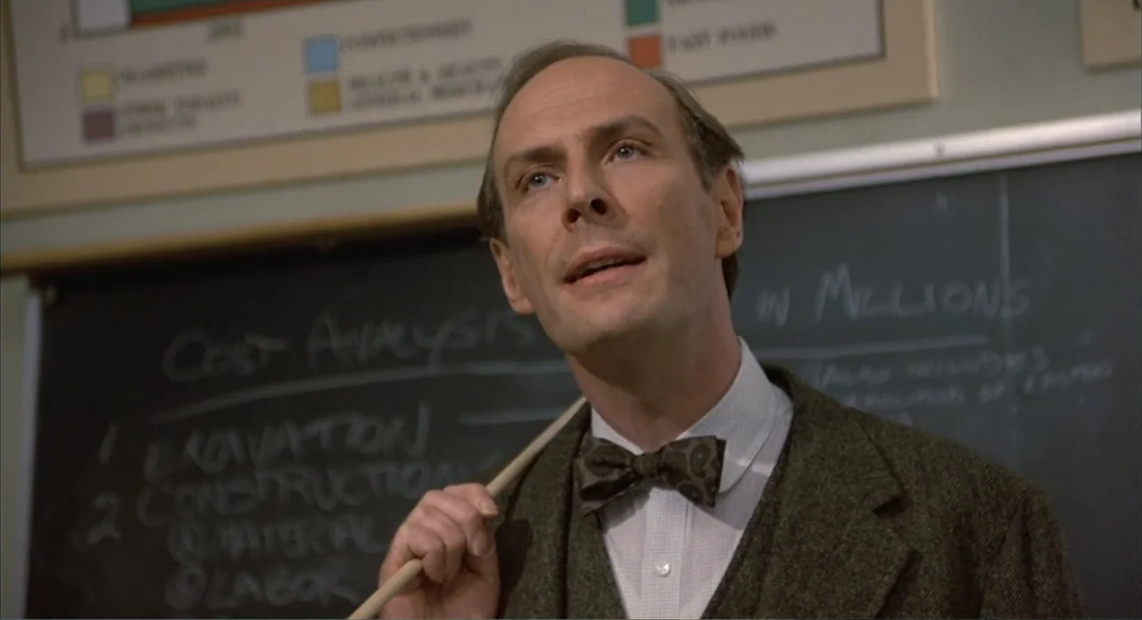 Back to School (1986) movie still: medium — A classroom features a black chalkboard covered in handwritten cost analysis notes. A middle-aged man in…