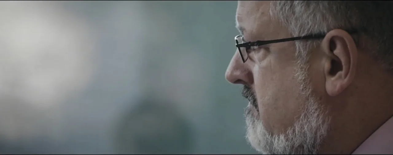 Hunting for Hedonia (2019) movie still: closeup — A man stands indoors against a muted gray wall background. The subject is in profile, gazing…