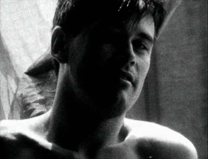 Glitterbug (1994) movie still: closeup — A dimly lit room features a thin white curtain near a window. A shirtless young man turns his head while…