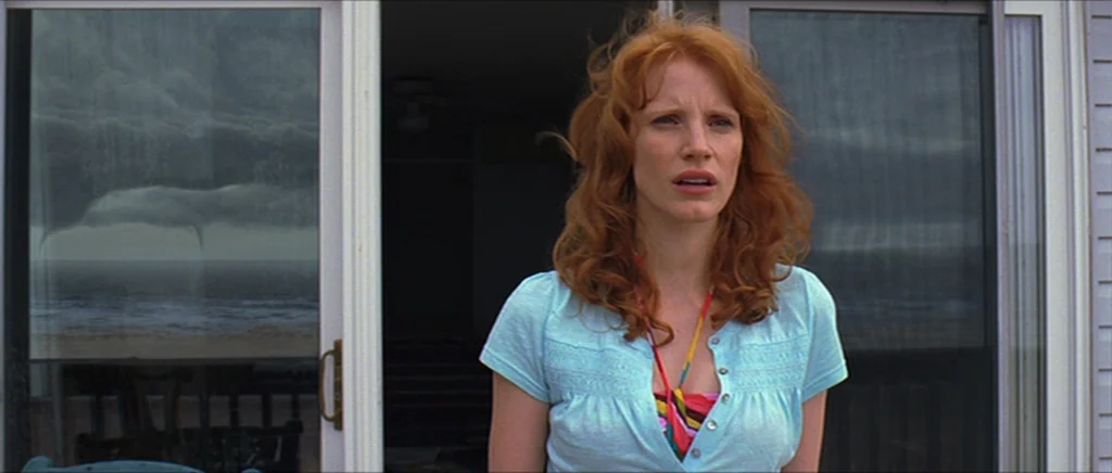 Take Shelter (2011) movie still: medium — A woman stands in the threshold of a beach house with glass sliding doors. Outside, dark storm clouds and…