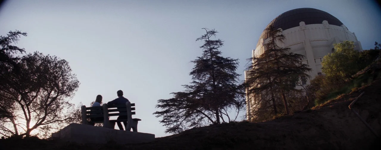 La La Land (2016) movie still: wide — A man and woman sit on a wooden bench overlooking a large domed observatory. The sky is clear during golden…