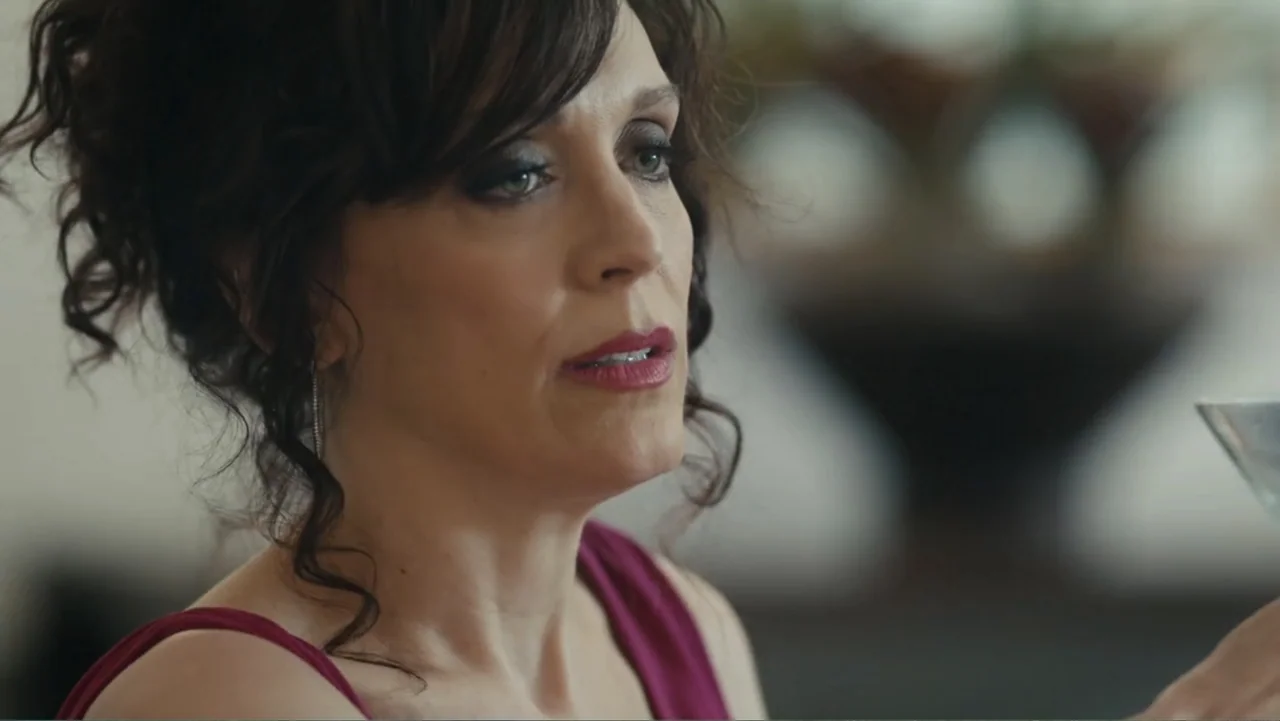 Toss It (2019) movie still: closeup — An interior living room features a person in a magenta dress in soft daylight. The subject holds a glass while…