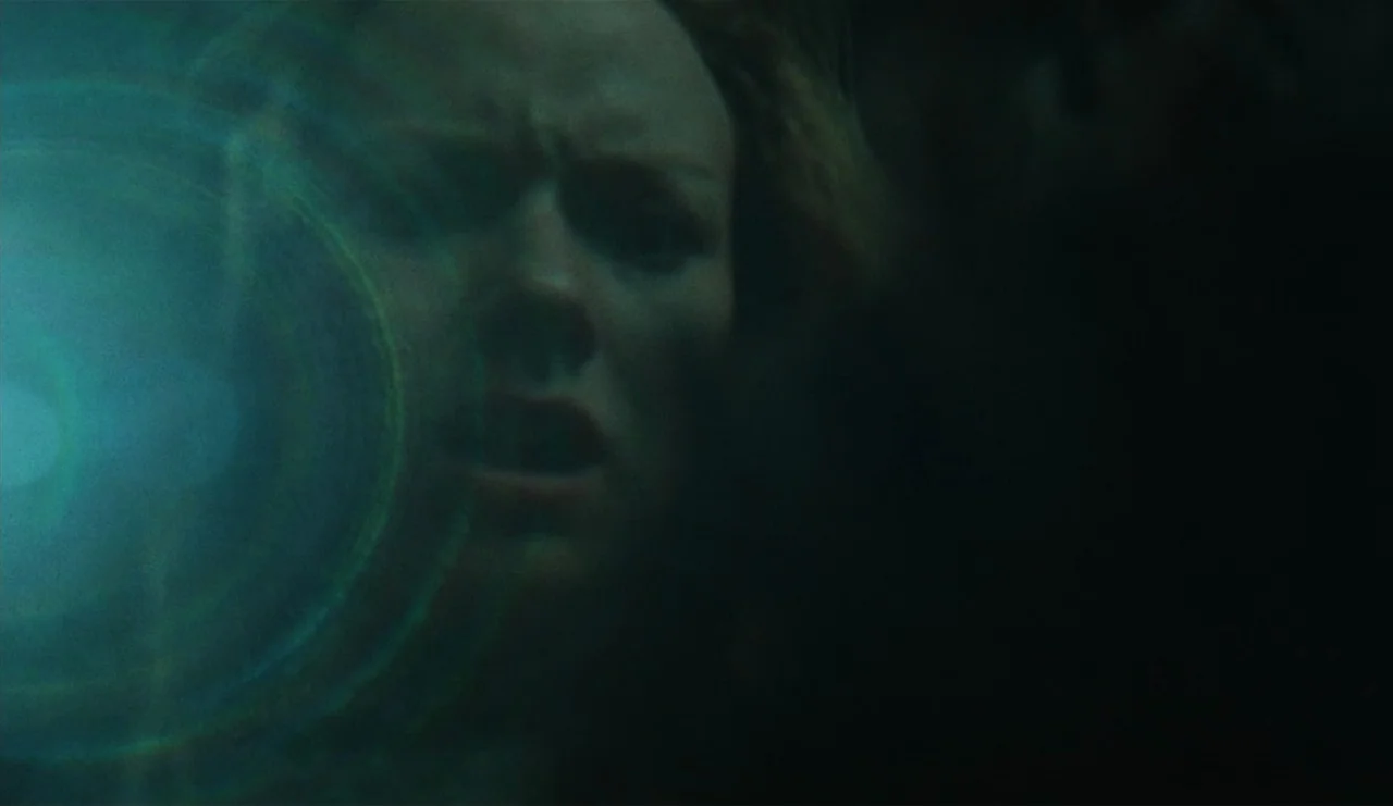 The Ring (2002) movie still: closeup — A woman's face is dimly lit in a dark, indistinct environment. A green lens flare obstructs part of the frame…
