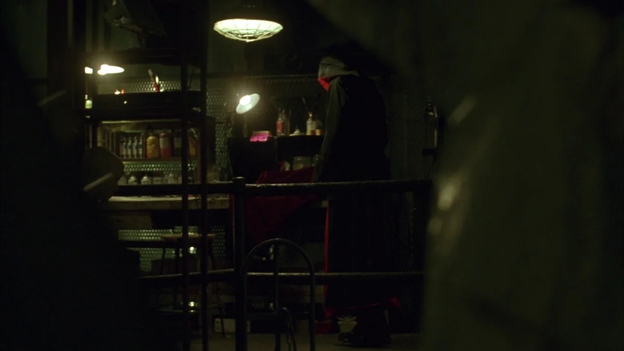 Saw movie still: medium — A dark, industrial workspace contains a workbench illuminated by multiple low-hanging pendant lights. A hooded person in…