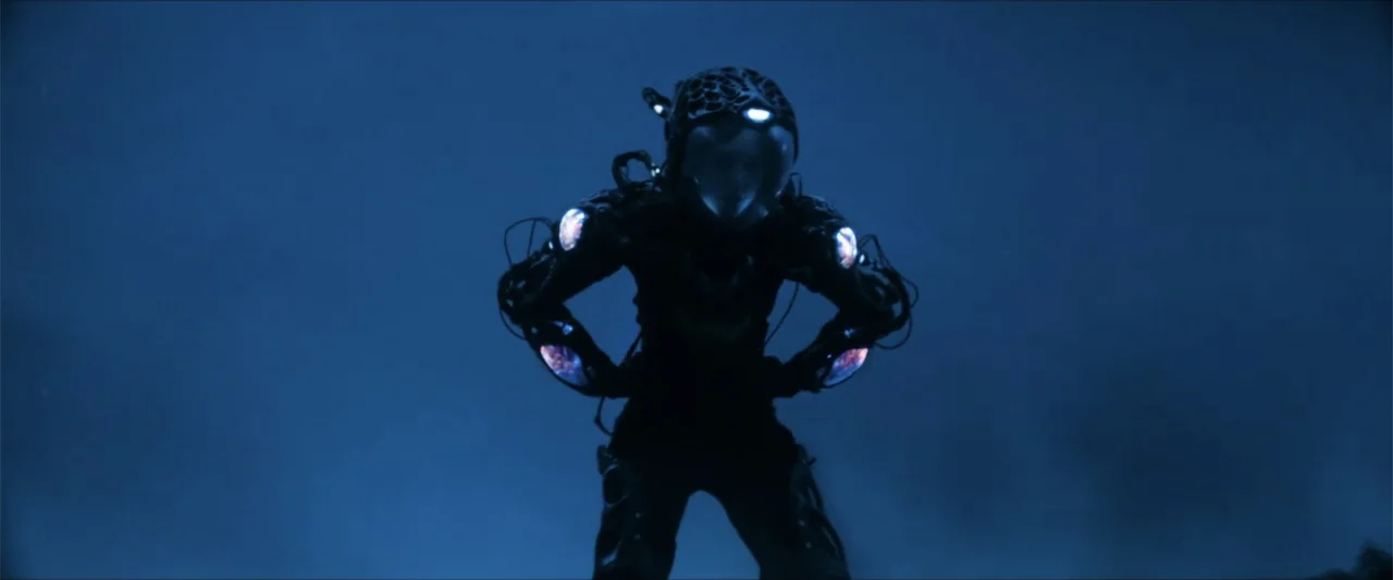 Ash (2025) movie still: fullbody — A dark, featureless environment serves as the backdrop for a futuristic, armored figure. The character wears a…
