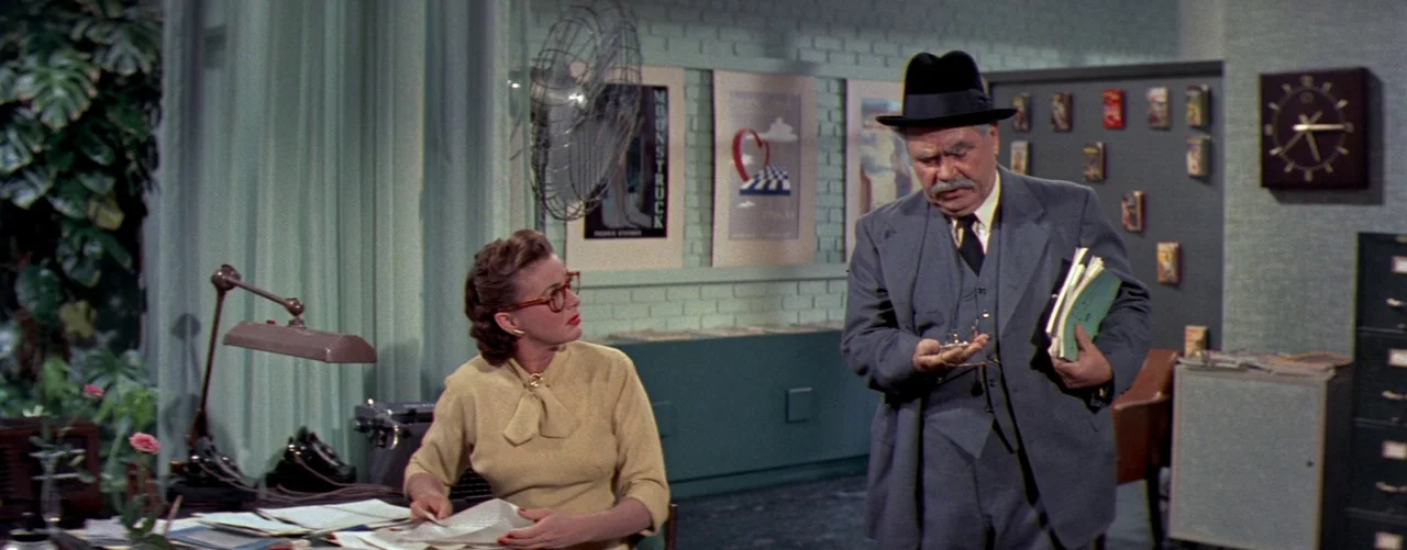 The Seven Year Itch (1955) movie still: medium — A light mint-green walled space decorated with framed posters and a wall-mounted clock. An elderly…