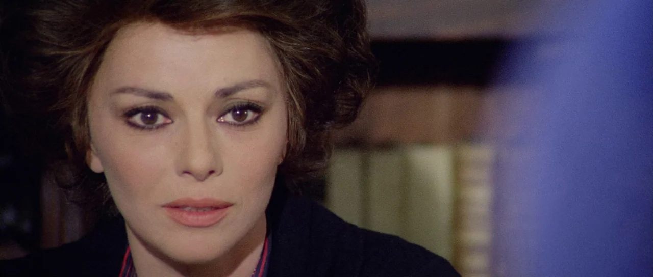What Have They Done To Your Daughters? (1974) movie still: closeup — A woman sits inside a room with shelves of books visible in the background. She…