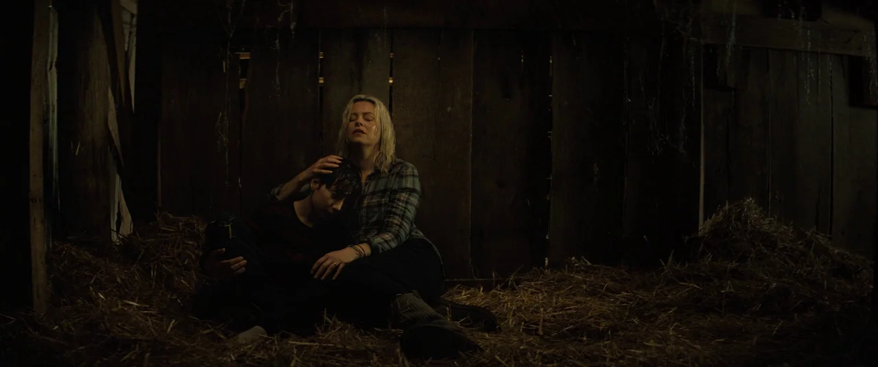 Brightburn (2019) movie still: medium — A dark wooden barn interior is filled with loose hay and scattered cobwebs. A woman sits on the ground with…