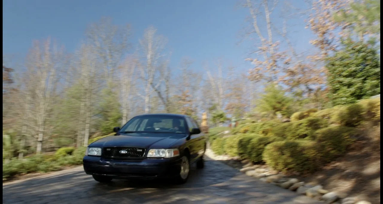 Sabotage (2014) movie still: wide — A dark sedan travels along a stone driveway surrounded by manicured shrubs and trees. The vehicle moves forward…