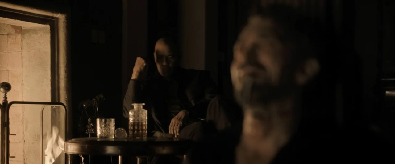The Equalizer 3 (2023) movie still: medium — A dimly lit room features a glowing fireplace next to a small wooden table. A man in the background…