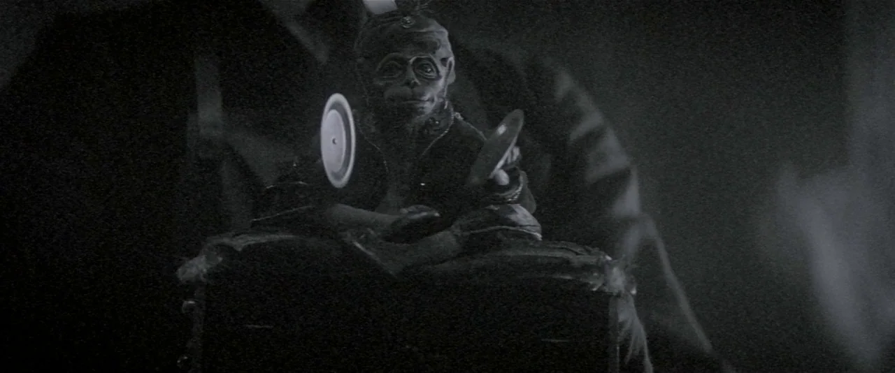 The Phantom of the Opera (2004) movie still: medium — A wind-up cymbal-banging monkey toy sits centered in a dark, shadow-filled environment. The…