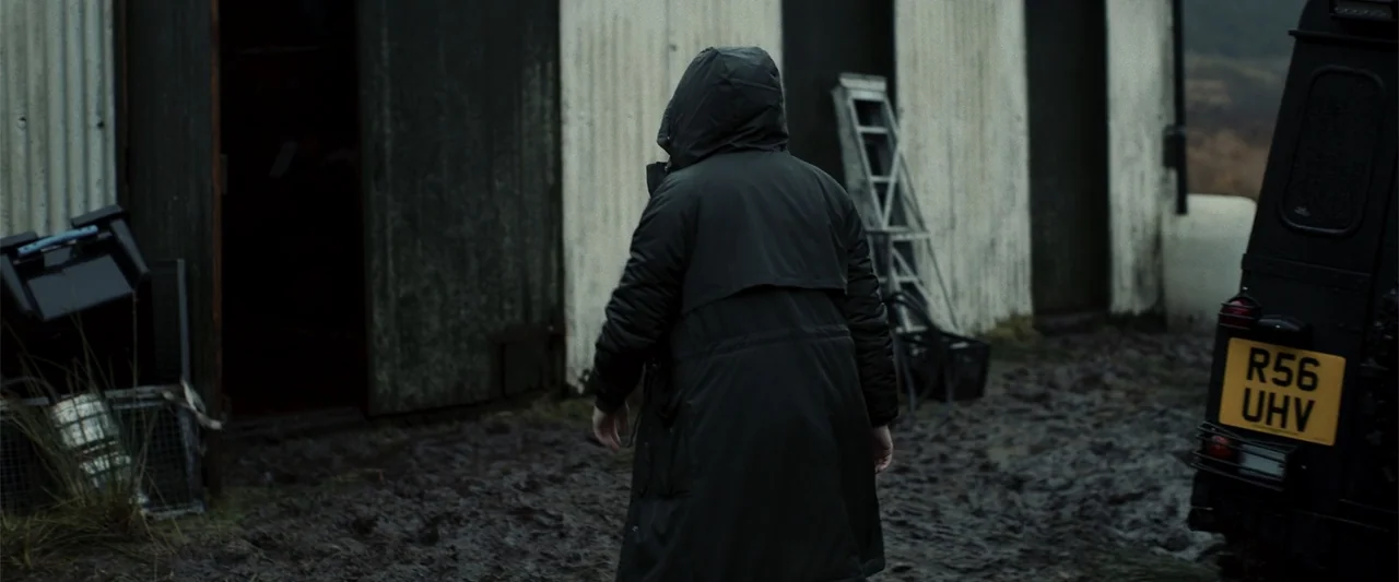 My Son (2021) movie still: medium — A muddy farmyard sits before a white corrugated metal shed on an overcast day. A person in a black hooded coat…