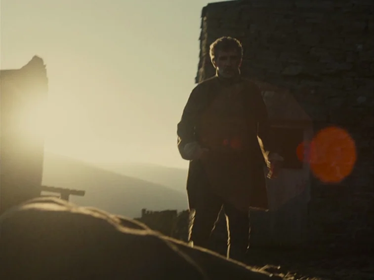 Wuthering Heights movie still: medium — A man stands outdoors in a rocky, barren landscape during sunset. He faces the camera while backlight from…