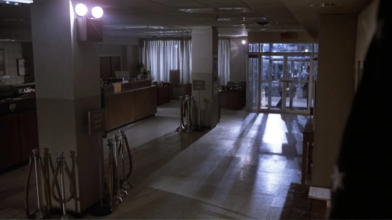 Dog Day Afternoon (1975) movie still: establishing — A quiet building lobby with a service counter and glass entry doors at night. The interior is…