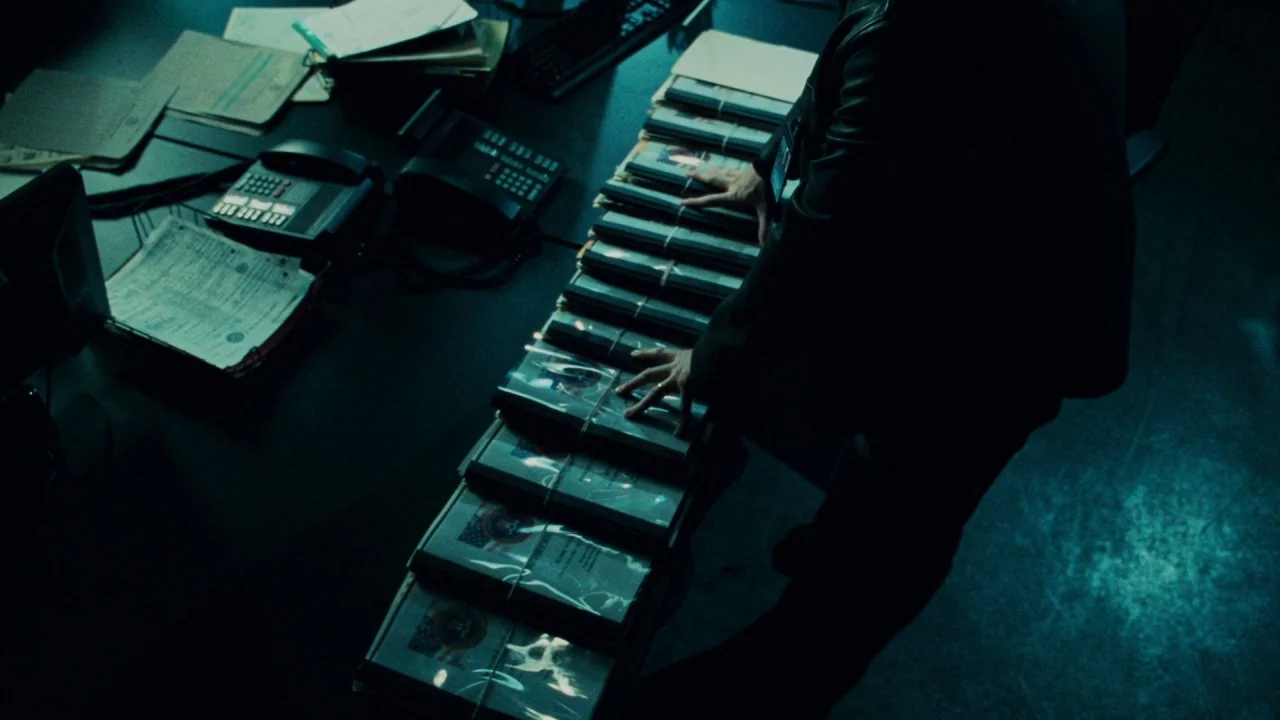 Saw V (2008) movie still: medium — A desk in a dark room holds a line of documents bound with rubber bands. A person stands facing the desk and…