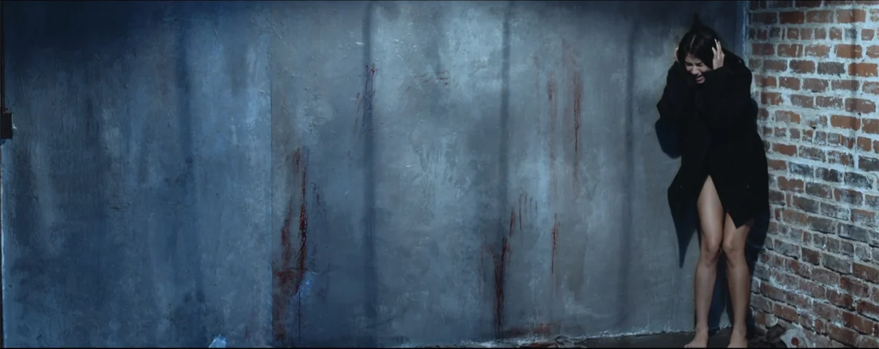 El Último Juego (2025) movie still: wide — A dark corner of a room features a grey concrete wall beside exposed brick. A woman stands in the corner…